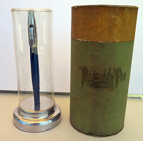 #4587: REYNOLDS INTERNATIONAL BALLPOINT IN DARK BLUE WITH ALUMINUM CAP WITH CIRCUMFERENTIAL GROVES. THE CAP REMOVES AND SNAPS ON OVER THE FRONT END OF THE PEN. ELABORATE DISPLAY CASE IN OLIVE GREEN. IT CONTAINS A VERY CLEAR CELLOPHANE COVER AND AN ALUMINUM BASE